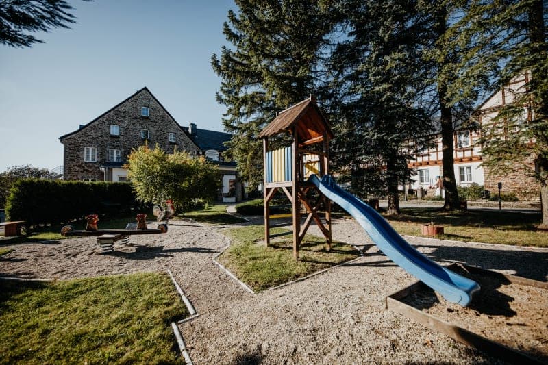 Kinderspielplatz mit blauer Rutsche und Schaukeltieren im sonnigen Hotelgarten vor historischem Steingebäude