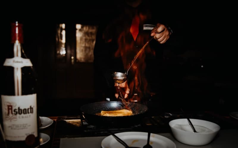 Koch flambiert Crêpes in einer Pfanne mit hohen Flammen, im Hintergrund eine Flasche Asbach