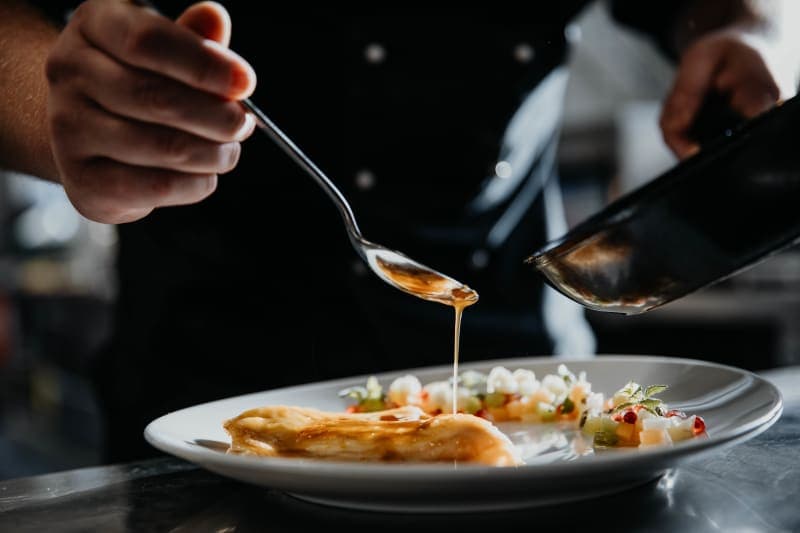 Koch gießt goldene Sauce mit einem Löffel über ein elegant angerichtetes Gericht auf weißem Teller