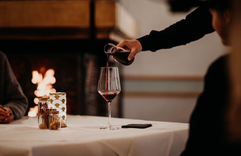 Rotwein wird in ein Weinglas eingeschenkt, im Hintergrund ein brennendes Kaminfeuer